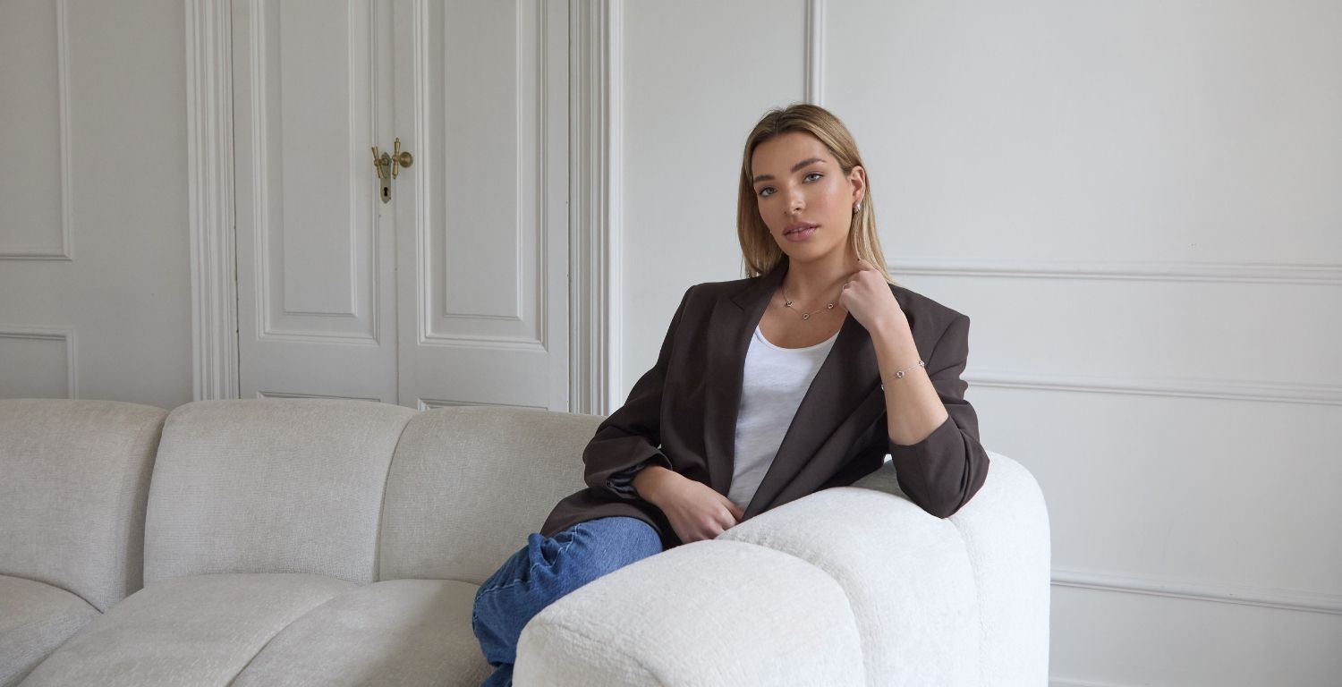 Woman. sitting on a white sofa in a room with light-coloured walls, wearing matching necklace and bracelet set jewellery