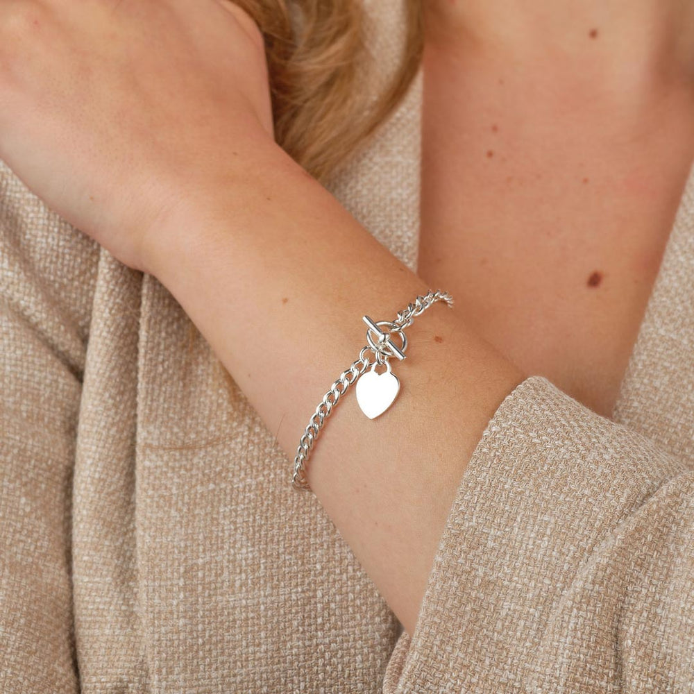 A close-up shot of a silver T-Bar bracelet with heart charm, made from polished silver-tone metal with a high-shine finish, featuring chunky chain links and a sleek T-Bar fastening, presented on a model
