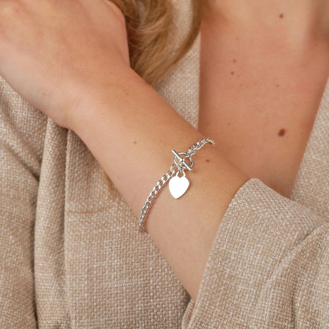 A close-up shot of a silver T-Bar bracelet with heart charm, made from polished silver-tone metal with a high-shine finish, featuring chunky chain links and a sleek T-Bar fastening, presented on a model