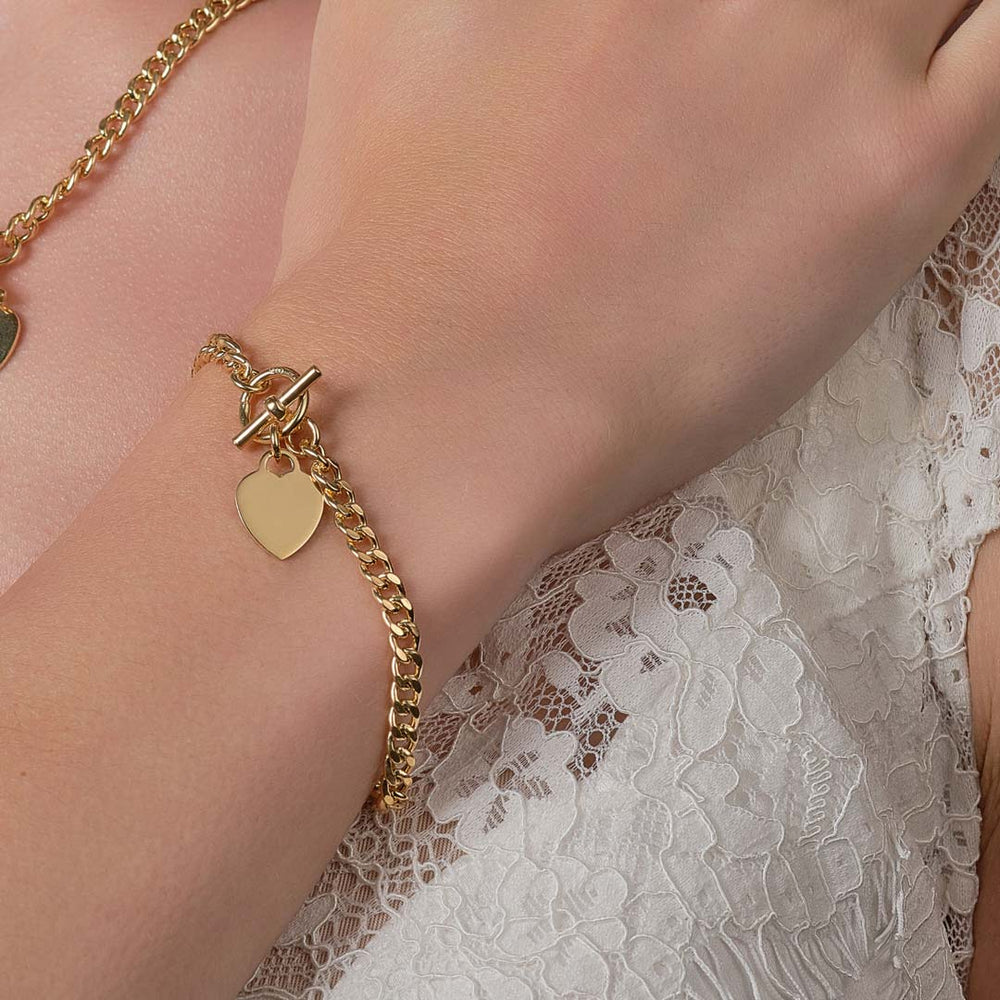 This image features a close-up lifestyle shot of a polished gold T-bar chain bracelet with a T-bar toggle clasp and a dangling heart charm, worn on a wrist against a white floral lace top.